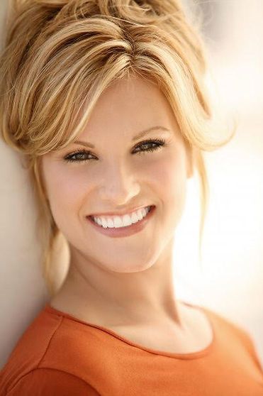 Woman with blonde hair, orange shirt, smiling against a light-colored wall.