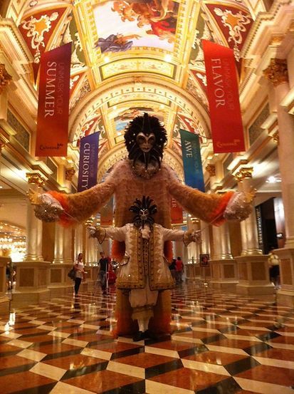 Masked performers in ornate costumes at the Venetian Hotel, with colorful banners and checkerboard floor.