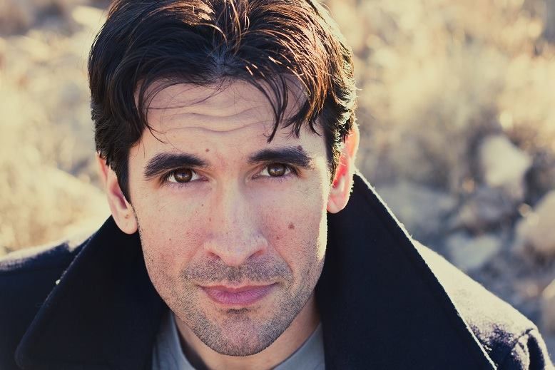 Man in a dark coat, looking towards the camera with a neutral expression, outdoor background.