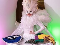 Drag queen in white feathered costume, mask, and headdress. Table with accessories in purple, yellow, and blue.