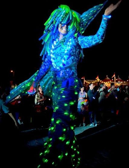 A person in a blue and green glowing costume on stilts, dancing at night.