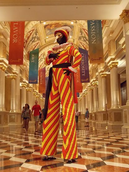 Stilt walker in colorful striped costume, inside a grand building with columns and banners.