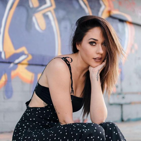 Woman with brown hair and a black outfit poses in front of a colorful graffiti wall.