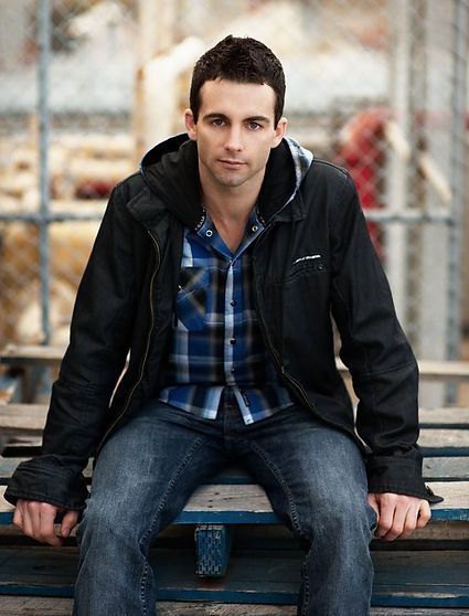 Man in a black jacket, blue plaid shirt, and jeans, sitting outdoors in front of a construction site.