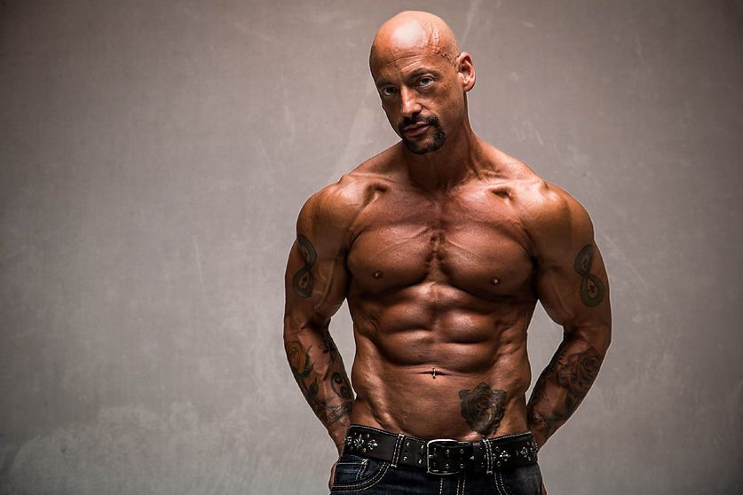 Bald muscular man with tattoos, hands in pockets, against a gray backdrop.