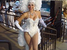 Woman in white showgirl outfit, feather headdress, smiling, standing near railing.