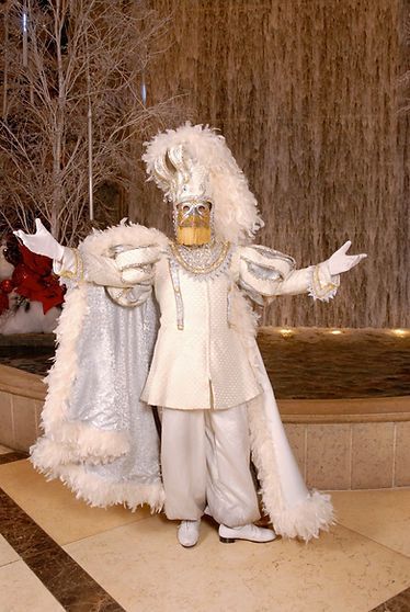 Person in elaborate white and gold costume with arms outstretched, standing near a water feature.