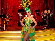 Woman in elaborate green costume, headdress, and stage setting, arms at her sides, smiles.