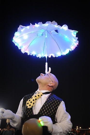 Man balances a lit, ruffled umbrella on his nose at night, wearing a vest and polka-dot tie.