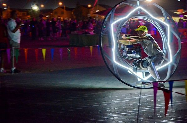 Person in a glowing orb performs on a dark street, surrounded by a crowd, wearing a neon green cap.