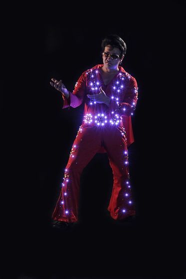Man in red Elvis jumpsuit, lit with purple lights, performing on dark stage.