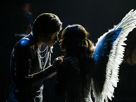 Two people in costume; one with angel wings, looking at each other in a theatrical setting.