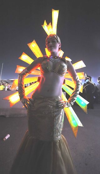 Woman in gold costume with glowing light display, arms akimbo, outdoor setting.