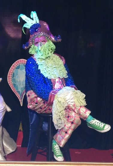 Person in colorful costume with light-up hat and beard, seated on a stool, wearing green sneakers.