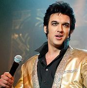 Man dressed as Elvis, wearing gold jacket, holding microphone, performing onstage.