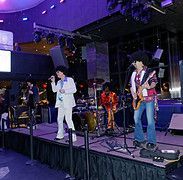 Band on stage performing, wearing wigs, playing instruments, in a venue with stage lights.