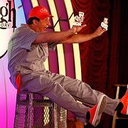 Magician seated, in red cap and shoes, juggling playing cards; performing on stage.