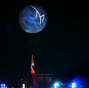 Aerial performer hanging from a red fabric, beneath a large, illuminated blue orb with a white logo, at night.