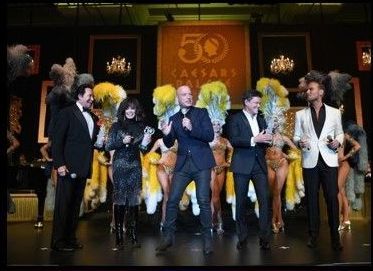 Men and women on stage at Caesars Palace 50th anniversary, showgirls in background.