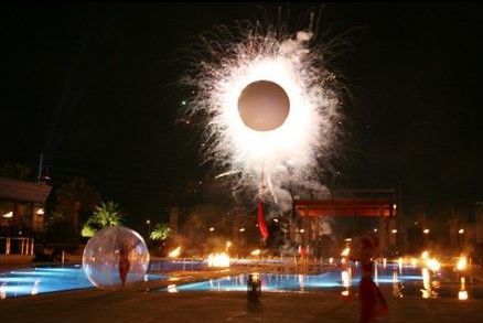 Fireworks burst around a black circle, possibly a balloon. Pool and torches in a night setting.