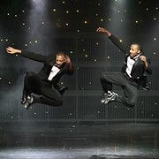 Two male dancers in tuxedos leaping in the air on a stage with a star-studded backdrop.
