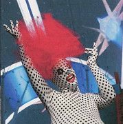 Person in polka-dot body suit with red wig, arms raised, smiling, in front of a colorful mural.