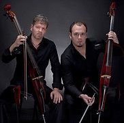 Two men, each with a red electric cello, wearing black. Studio shot, dark background.