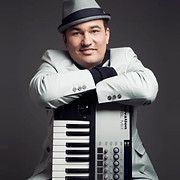 Man in a grey suit and fedora leans on a keyboard, smiling.