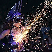 Woman in futuristic costume, sparks flying from a grinder. Dark background.