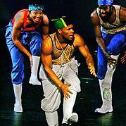 Three male dancers in colorful outfits, posing with athletic stances on a stage.