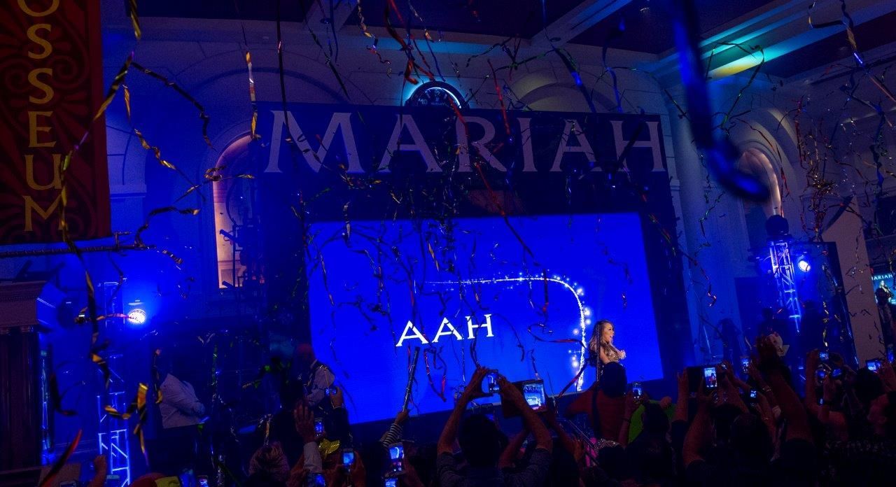 Mariah Carey performing onstage, blue lights, confetti falling. Crowd cheering with phones up.