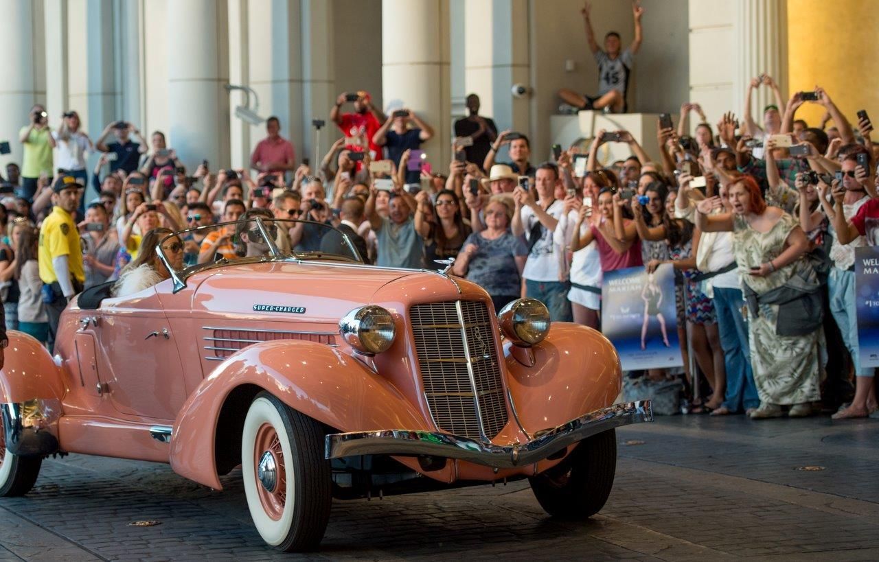 Pink vintage car driving through a crowd of people taking photos.