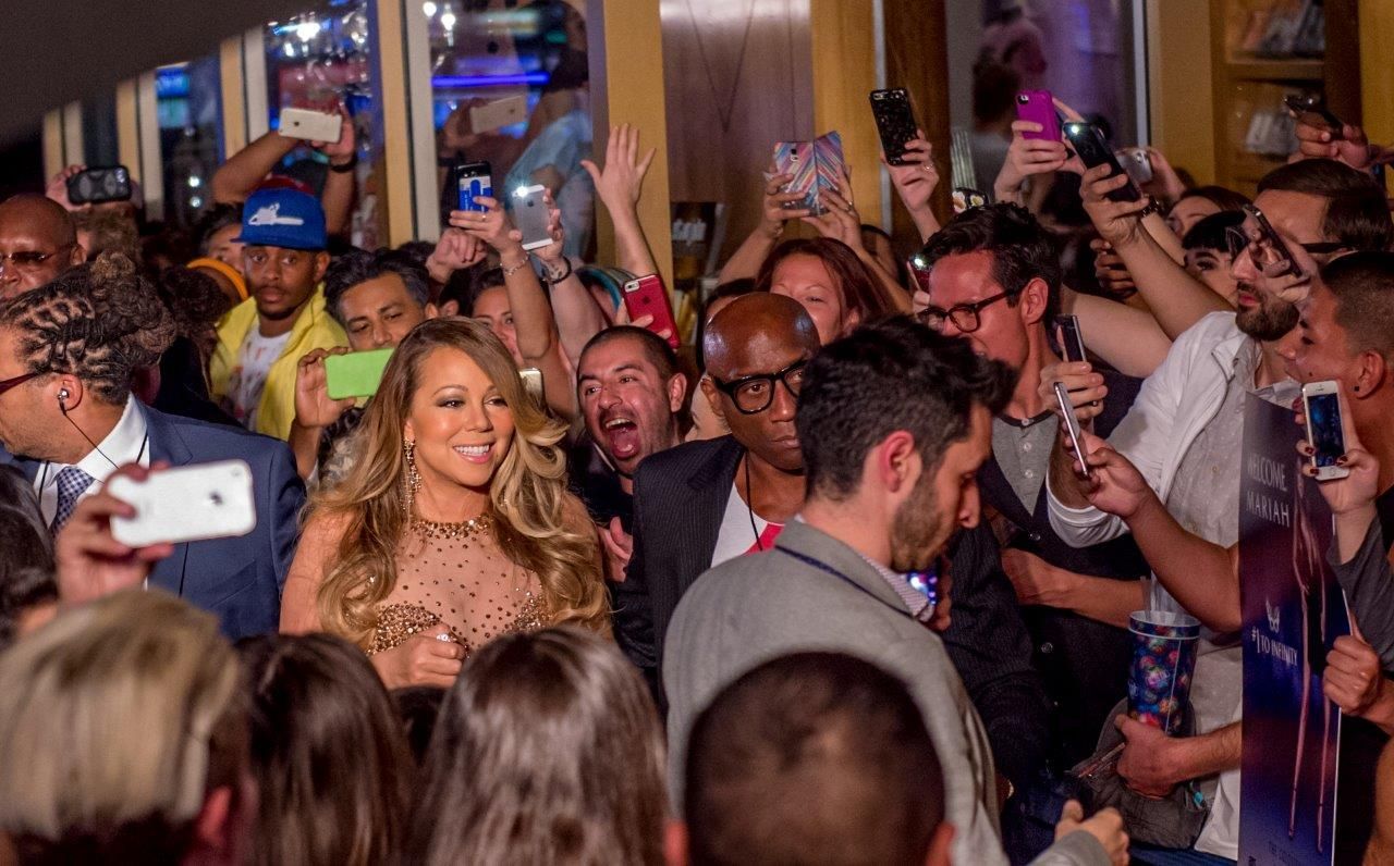 Mariah Carey smiles, surrounded by fans taking photos at an event.