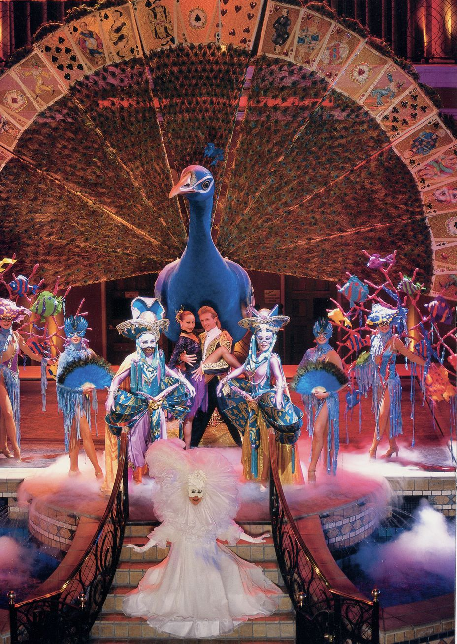 Stage show with a large blue peacock statue and performers in colorful costumes.