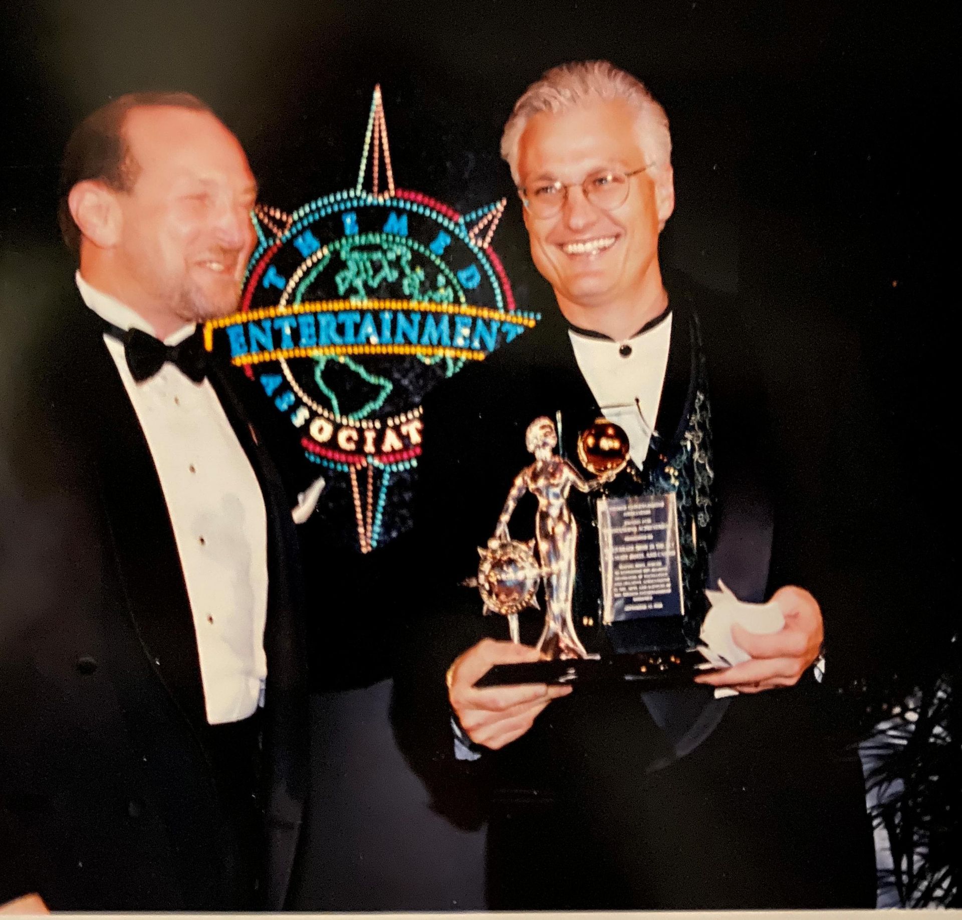 Two men at awards ceremony. One holds trophy, smiles. Logo in background,