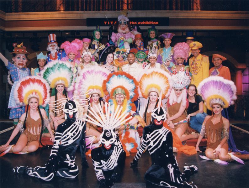 A large group of costumed performers poses on a stage. Variety of colorful headdresses and costumes.
