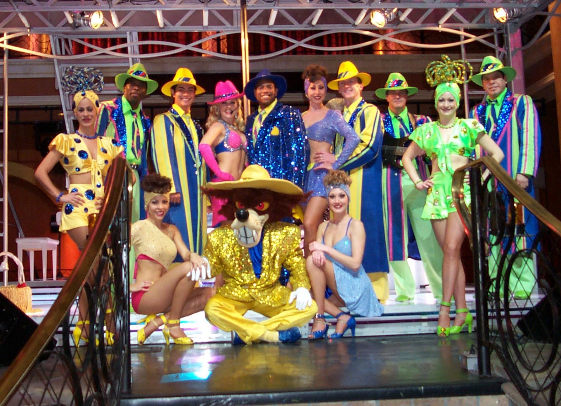 A group of costumed performers on a stage. Bright colors: yellow, blue, and green. A lion in a gold suit kneels.