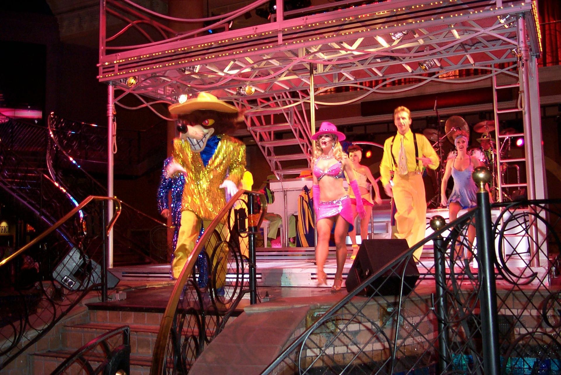 Performers in colorful costumes on a stage with lights; a man in gold, a woman in pink, others dancing.