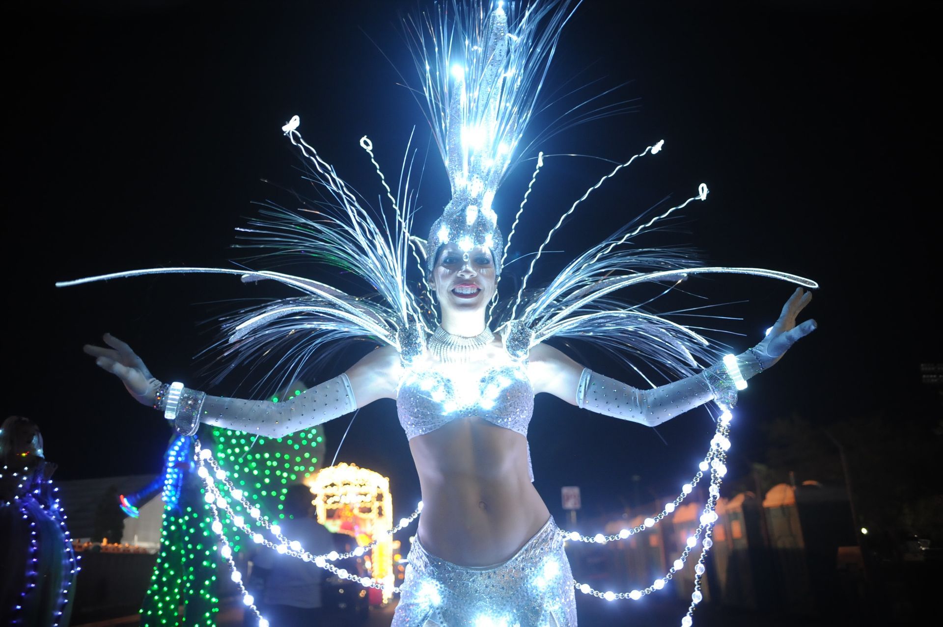 Woman in glowing white costume, arms outstretched, smiling. Nighttime outdoor performance.