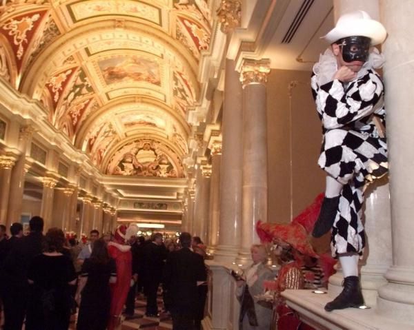 A person in harlequin costume with black mask poses by a pillar in a decorated Venetian style hallway.