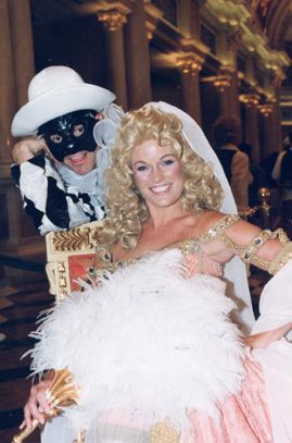 Woman in a blonde wig and gown smiles, with a man in a harlequin mask at her side, in a large hall.