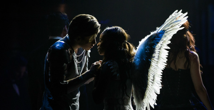 Couple in silhouette, illuminated by angel wings. One is wearing wings, other touches face. Stage.
