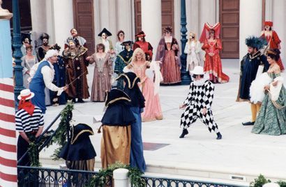 A group of people in period costumes on a stage; a harlequin dances in the foreground.