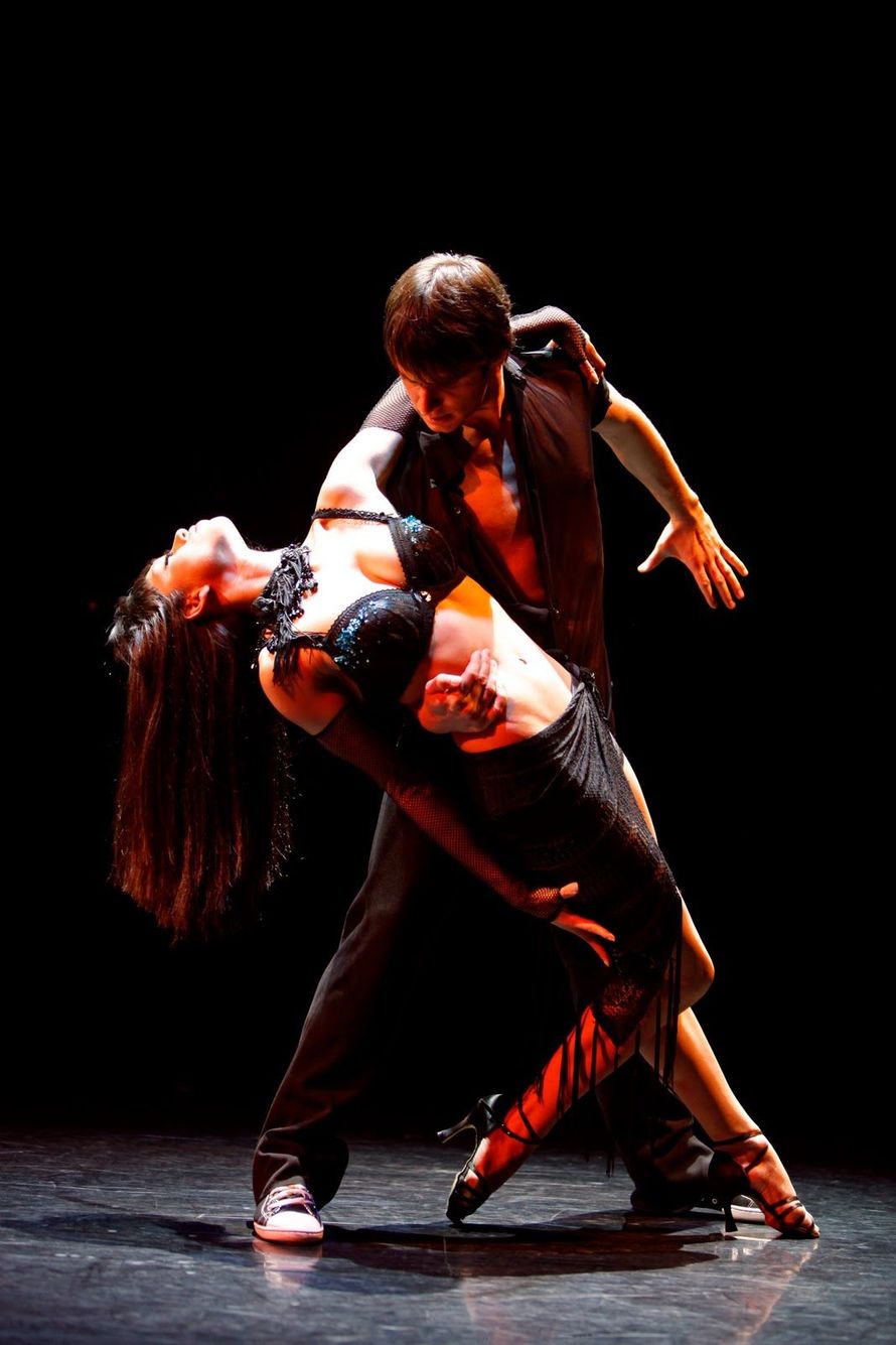 Dancers in a passionate tango pose. Woman in black dress leans back as man supports, both on a dark stage.
