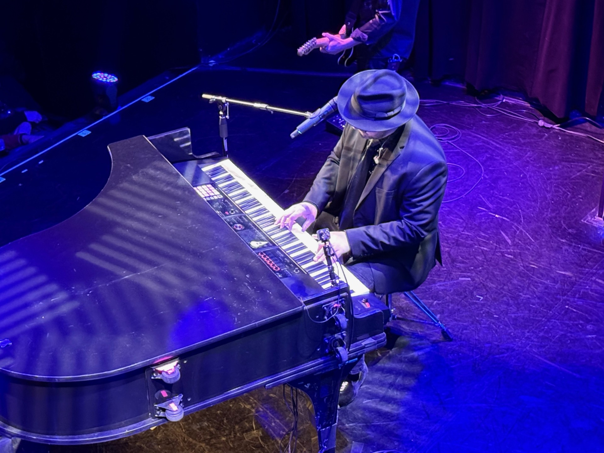 Pianist in a blue suit and fedora performing on stage, illuminated by stage lights.