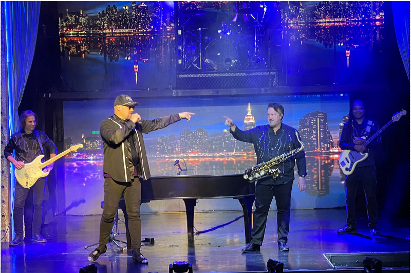 Band performing on stage, man singing, another playing saxophone. New York City skyline backdrop. Blue lighting.