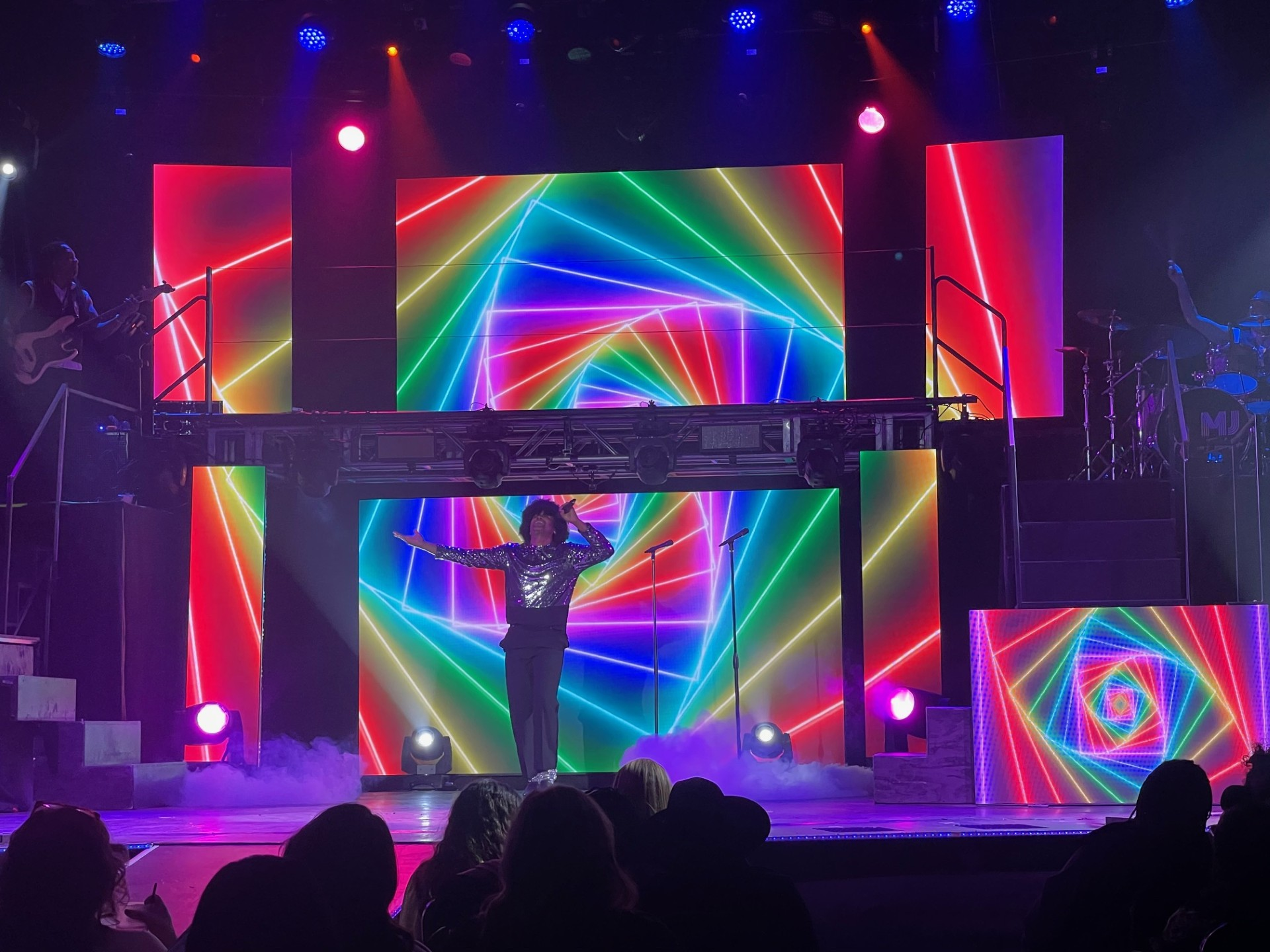 A performer on stage with rainbow-colored LED panels, arms outstretched. Band on left.