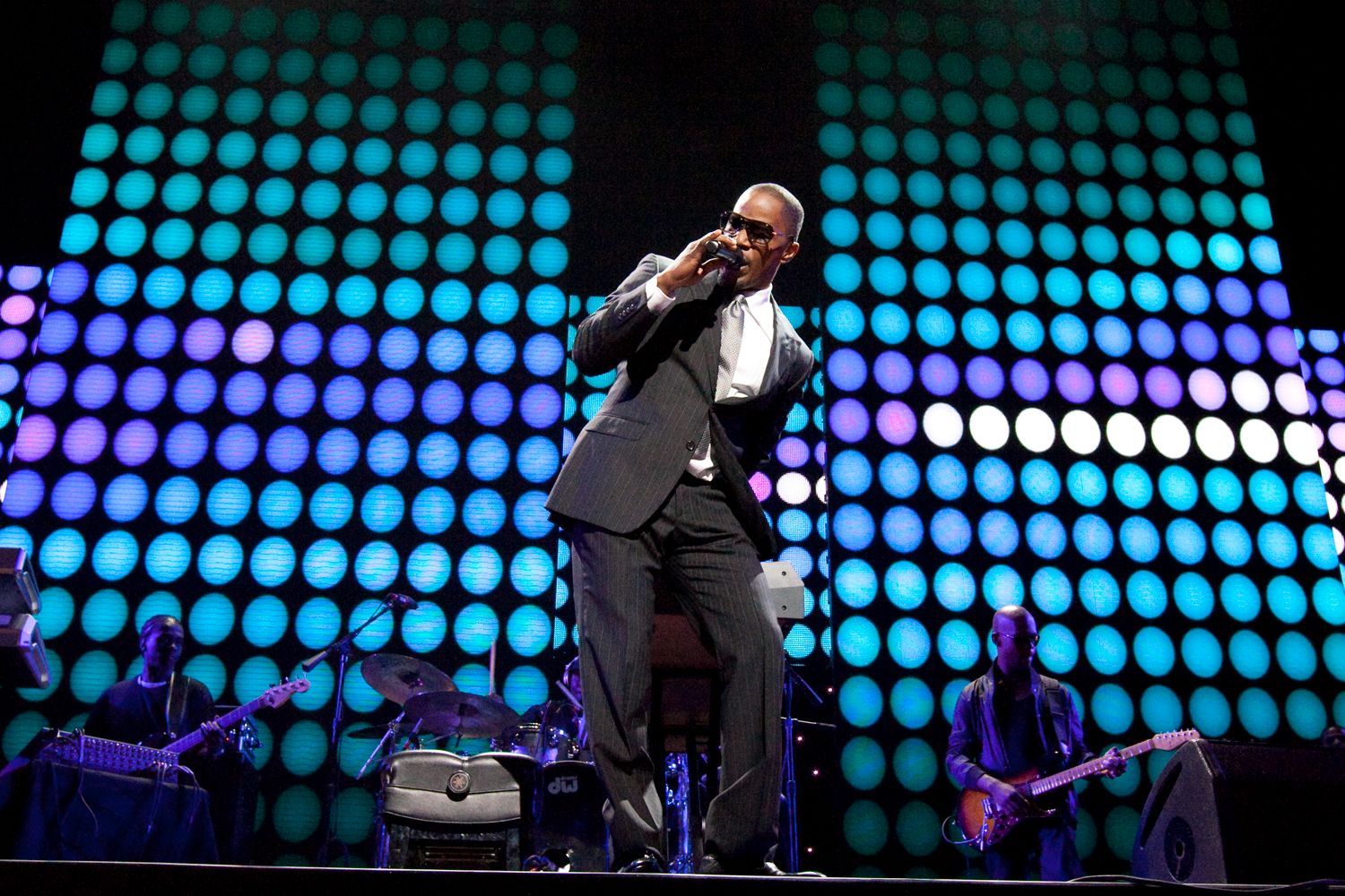 Singer in a suit performs on stage with a band, under a colorful LED light display.