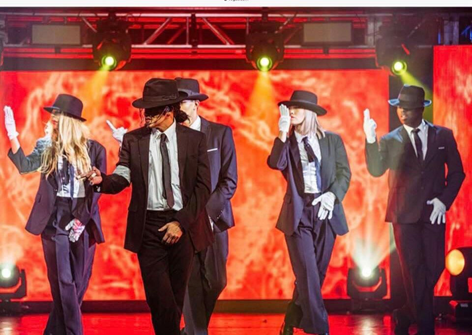 Dancers in suits, hats, and white gloves perform on stage with red lighting.