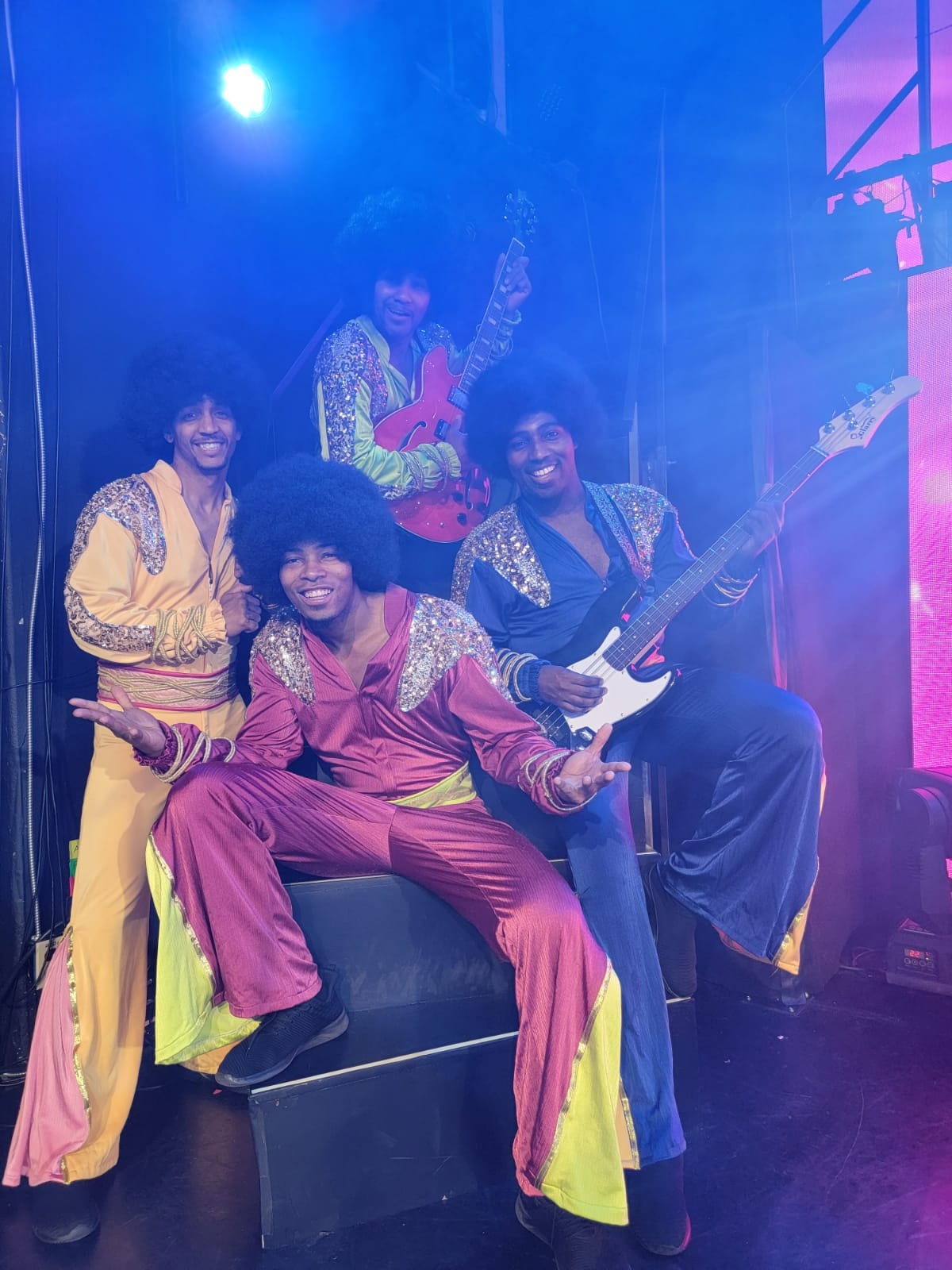 Four people in 70s-style outfits with afros. They are playing instruments on a stage with blue lighting.